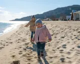 Dwa uśmiechnięte dzieci biegają po piaszczystej plaży. Słońce odbija się od wody, a w tle widać wzgórza i budynek.