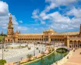 Der beeindruckende Sevilla Palast mit seinen Türmen und Arkaden, umgeben von einem Kanal und belebtem Platz mit Brunnen und Kutschen.