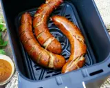 Drei frische grobe Bratwürste garen in einem Airfryer. Daneben eine Schale mit Dip und Petersilie.