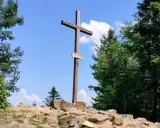 Drewniany krzyż na skalistym szczycie w Górach Świętokrzyskich. Otaczają go bujne drzewa i błękitne niebo z chmurami.