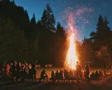 Människor samlas runt en stor brasa i skogen, en vacker svensk tradition som lyser upp natten med gnistor och värme.