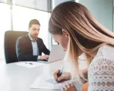 Kobieta podpisuje dokumenty, planując jak zmienić nazwisko po rozwodzie. Mężczyzna w tle pracuje przy laptopie.