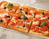 Chrupiąca pizza z ciasta francuskiego z salami, pomidorkami i oliwkami, udekorowana listkami bazylii.