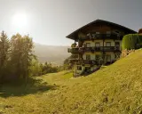 Haus im Alpenvorland mit traditioneller Architektur und Balkonen, umgeben von grünen Wiesen und Bergen.