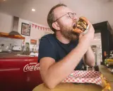 Mężczyzna z brodą i okularami zajada ogromnego burgera. W tle czerwona lodówka Coca-Coli, która może być jedną z ciekawostek o USA.