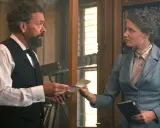 En scen från en alfred nobel film. En man med skägg ger ett brev till en kvinna i en blå dräkt framför en bokhylla.
