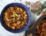 Un reconfortante plato de pasta e fagioli en un cuenco azul, con frijoles y romero esparcidos alrededor.