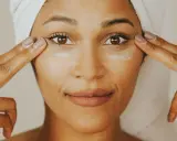 Une femme applique de la crème contour des yeux sur tout le visage, avec une serviette sur la tête.