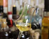 Ein Glas mit goldgelbem Wein steht vor einer Sammlung von Flaschen. Das Glas erinnert an ein Kirchenfenster, das Licht bricht.
