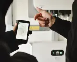 En person håller upp en mobiltelefon med sesol appen som visar solenergi-data. En annan person pekar mot en vit enhet i bakgrunden.