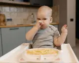 Niemowlę w krzesełku do karmienia próbuje samodzielnie jeść kaszkę z talerzyka.