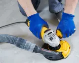 Ręce w niebieskich rękawicach trzymają szlifierkę z odkurzaczem, pokazując, jak wyszlifować beton.
