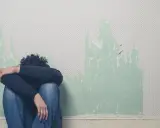 Osoba siedząca na podłodze, skulona, z głową ukrytą w ramionach. Tło to ściana z odpadającą tapetą, sugerująca trudne emocje, być może związane z depalin skutki uboczne.