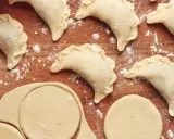 Gotowy do lepienia pierogów z idealnym, miękkim ciastem. Na drewnianej desce widać surowe pierożki i wykrojone kółka z ciasta.