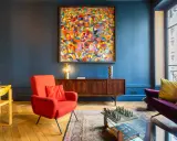 Ein Wohnzimmer mit einem farbenfrohen Gemälde im art déco zeitraum, rotem Sessel, Holzsideboard und einem Schachspiel auf dem Couchtisch.