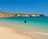 Ein Mann im Neoprenanzug steht im türkisfarbenen Wasser am Praia do Martinhal. Im Hintergrund ragen ockerfarbene Klippen auf.