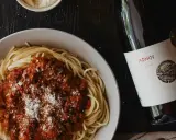 Plato de espaguetis con salsa boloñesa y queso rallado, acompañado de una botella de vino español Adnos.
