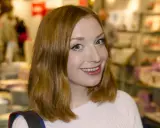 Sandra Beijer blogg: En glad kvinna med rött hår ler mot kameran i en ljusrosa tröja. Bakgrunden är suddig och visar en butiksmiljö.
