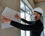 Specjalista BHP w białym kasku i masce analizuje projekt budowlany przyklejony do ściany.