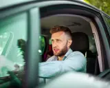 Mężczyzna z zarostem, w jasnej koszuli, siedzi za kierownicą. Jego profil sugeruje skupienie, jakby wypełniał profil kierowcy wniosek.