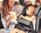 Uśmiechnięta mama z pluszowym zajączkiem przytula córkę w foteliku samochodowym Cybex Pallas 9-36 kg.