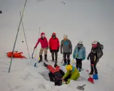 Grupa ratowników ćwiczy akcję po lawinie w Alpach. Jedna osoba jest zasypana, inni szukają jej z sondami i łopatami.