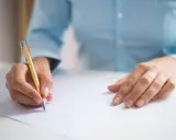 Dłoń pisze złotym długopisem na białej kartce, przygotowując się do egzaminu CAE practice tests.
