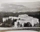 Duży budynek sanatorium w otoczeniu drzew, na tle pagórkowatego krajobrazu.