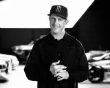 Ken Block, sonriente y con gorra, posa frente a coches de rally.