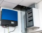 Niebieski falownik, kluczowy element instalacji fotowoltaicznej, zamontowany obok białej skrzynki z zabezpieczeniami.