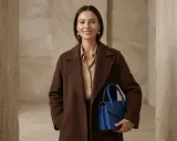 Elegancka kobieta w płaszczu w kolorze koniakowym trzyma niebieską torebkę.