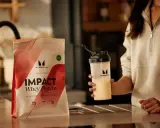 Eine Person hält einen Shaker mit einem cremigen Getränk. Daneben liegt eine Packung Myprotein Impact Whey Protein. Perfekt für den proteinshake vor dem schlafen.