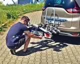 Mężczyzna montuje trzecią tablicę rejestracyjną WE 180SX na bagażniku rowerowym przy samochodzie.