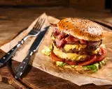 Soczysty burger z podwójną porcją mięsa, serem, bekonem i warzywami na desce z widelcem i nożem.