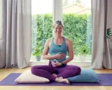 Kvinna i lila kläder sitter i meditationsställning på en yogamatta, med händerna över magen. Hon utövar yoga gravid.