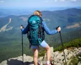 Kobieta z turkusowym plecakiem i kijkami trekkingowymi podziwia widok na góry. Idealny plecak w góry na wyprawę.