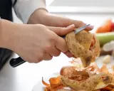 Ręce obierają ziemniaki, przygotowując je do gotowania. Obok leżą skórki, marchewki, papryka i czosnek. To część zwrotu artykułów spożywczych.