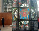 Un vitrail coloré, représentant des scènes bibliques, se dresse dans un intérieur. Un homme consulte son téléphone devant ce chef-d'œuvre.