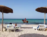 Piękna plaża na Djerbie, Tunezja. Jeździec na koniu przemierza brzeg morza, podczas gdy inni odpoczywają pod parasolami.