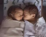 Dwa śpiące niemowlaki, każde w wieku około 2 miesięczne, wtulone w siebie pod białymi kocykami.