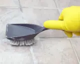 Rękawica w żółtej rękawiczce czyści szczotką i szufelką kafelki w kuchni z tłuszczu.