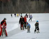 Människor åker norrviken skridskor på en isbelagd sjö. En pappa hjälper sitt barn, en hund dras på isen, och andra njuter av vintersporten.