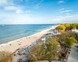 Słoneczna kołobrzeg plaża z ludźmi spacerującymi i odpoczywającymi. W tle morze i molo, a po prawej stronie drzewa w jesiennych barwach.