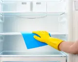 Ręka w żółtej rękawiczce czyści wnętrze lodówki niebieską gąbką. Dowiedz się, czym myć lodówkę, by była lśniąca i higieniczna.