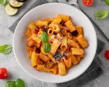 En skål med pasta alla norma, toppad med basilika och riven ost. Tomater och aubergineskivor ligger bredvid.