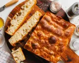 Goldbraune Focaccia mit grobem Salz, bereit zum Belegen. Daneben Olivenöl, Gewürze und ein Messer.