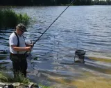 Wędkarz łowi ryby na jeziorze wierzbiczańskim. W wodzie stoi podbierak, a w tle widać zarośla i las.