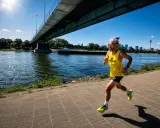 Mann joggt bei 30 Grad am Fluss entlang unter einer Brücke. Sonnenschein, Wasser und eine Skyline im Hintergrund.