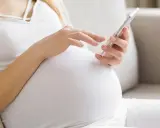 Kobieta w ciąży, w białej bluzce i kardiganie, siedzi na kanapie i korzysta z telefonu. Może sprawdzać zwolnienie lekarskie w ciąży.