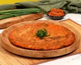 Ein goldbrauner, herzhafter Kimchi-Pfannkuchen, garniert mit Frühlingszwiebeln, liegt auf einem Holzteller. Daneben eine Schale mit Kimchi und grüne Frühlingszwiebeln.